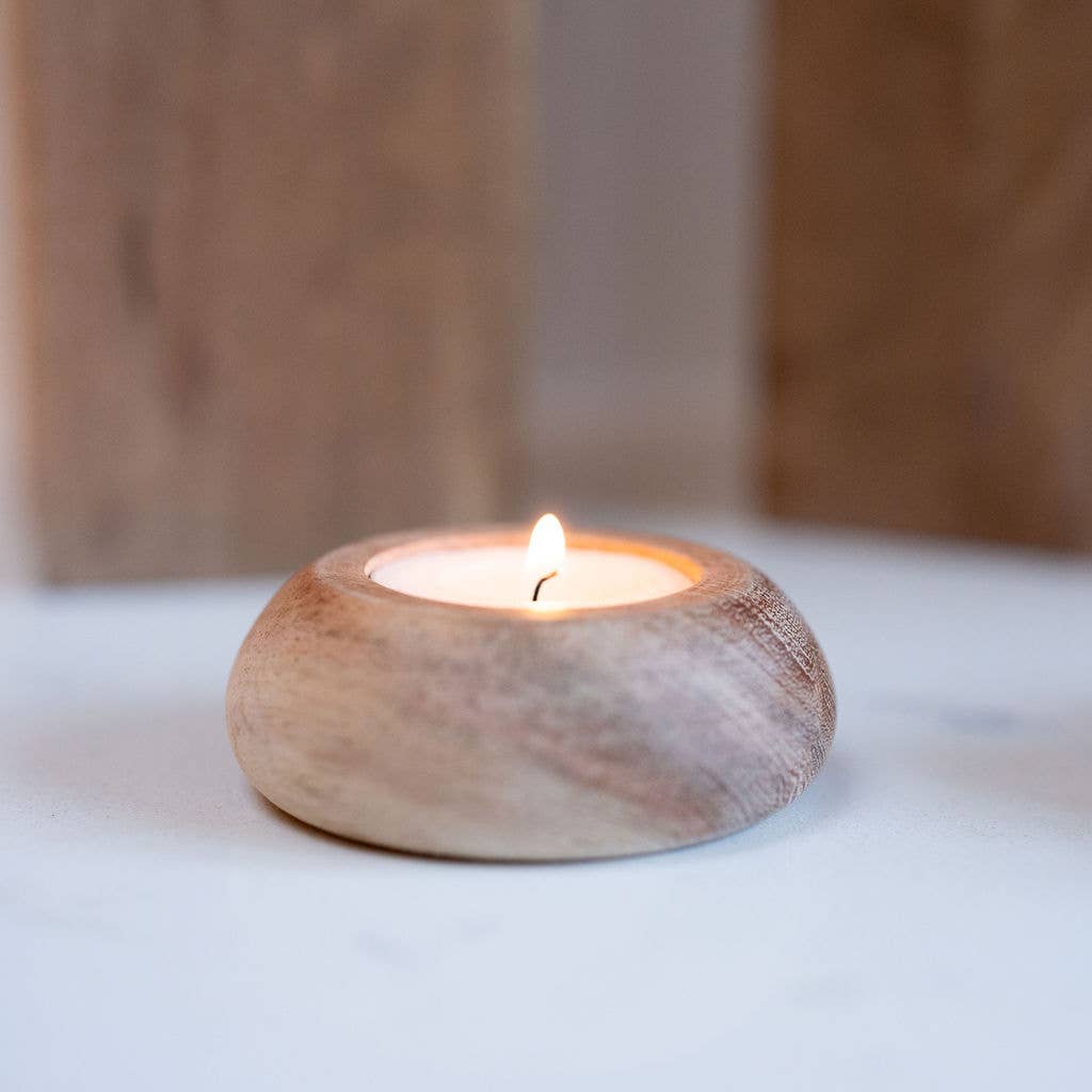 A small narbonne round wooden tea light holder with a lit tea light candle, set on a light surface with a softly blurred wooden background.