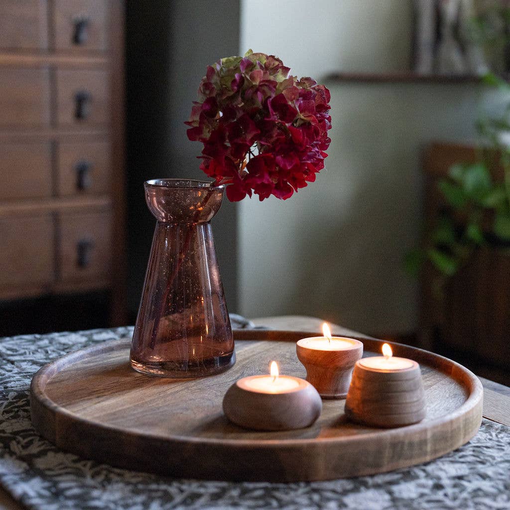 A styled tabletop scene with a round wooden tray holding three lit tealight candles in wooden tea light holders and a translucent amber glass vase with a dark red hydrangea bloom, set in a softly blurred indoor room.