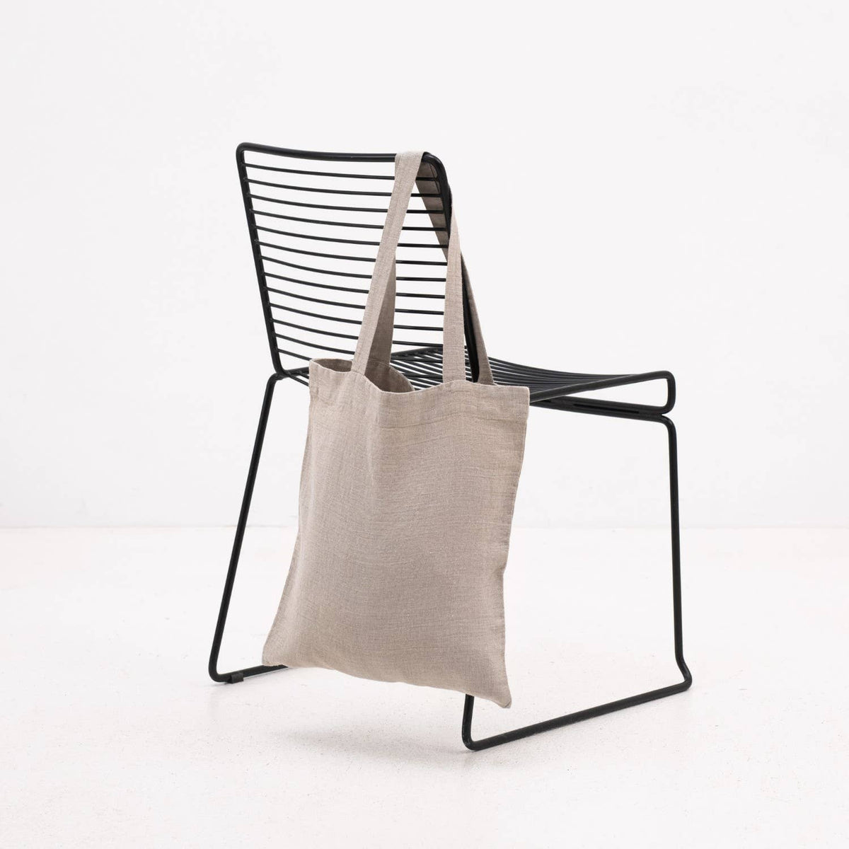 A natural linen tote bag resting on a black metal chair against a plain white background.