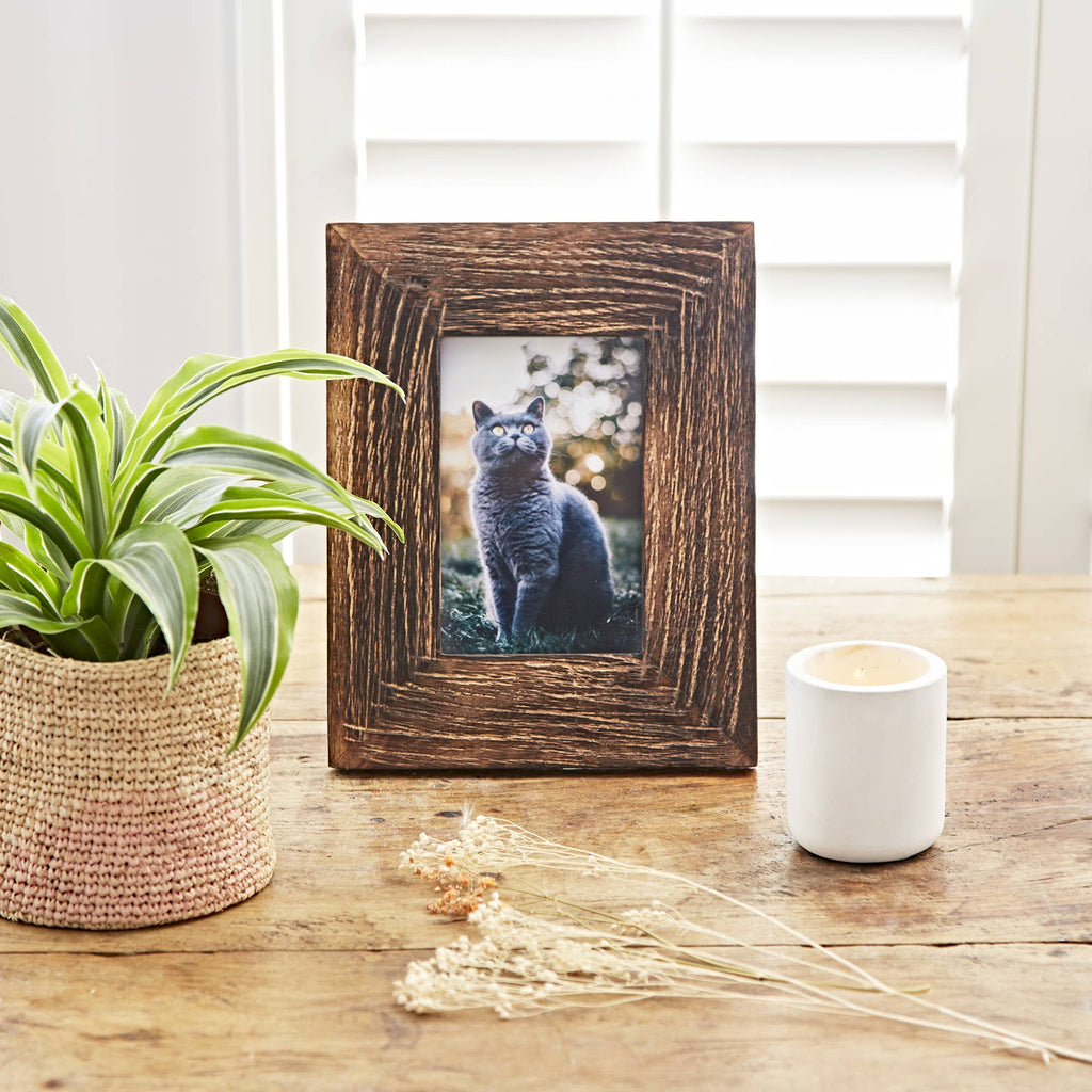 Rustic dark brown picture frame on a wooden table beside a potted plant and white candle holder.