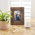 Rustic dark brown picture frame on a wooden table beside a potted plant and white candle holder.