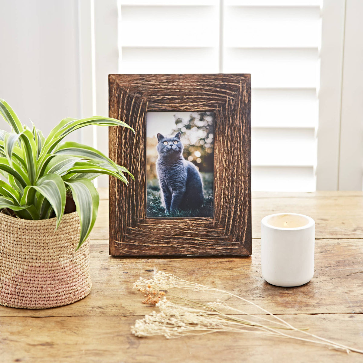Rustic dark brown picture frame on a wooden table beside a potted plant and white candle holder.