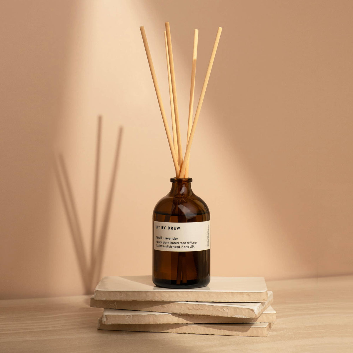 Amber glass reed diffuser with six natural wood reeds on stacked books against a warm beige wall.
