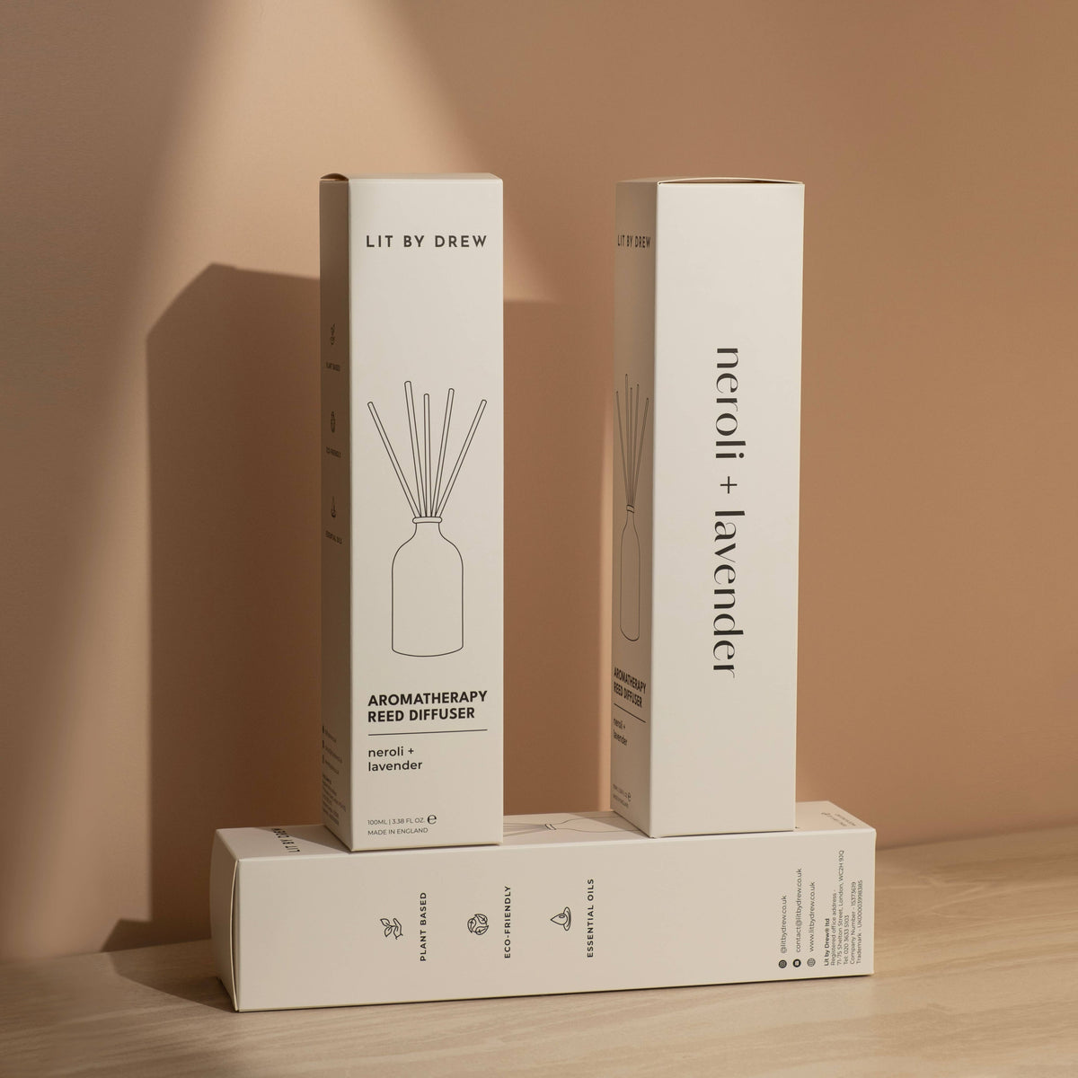 Three white Lit by Drew reed diffuser packaging boxes stacked on light wood against a beige wall.