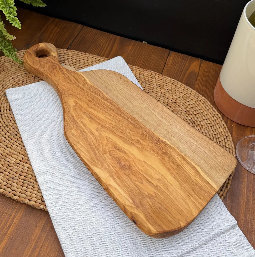 Large olive wood serving board with handle resting on a white cloth over a woven placemat.