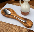 Olive wood spoon rest holding a stainless steel spoon on a white cloth over a woven placemat.