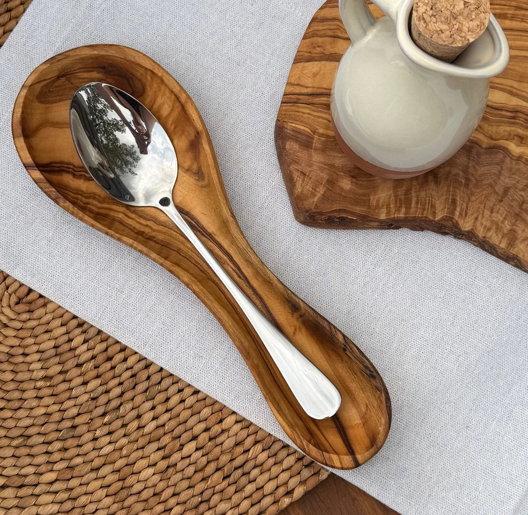 Olive wood spoon rest with a stainless steel spoon on a light gray cloth over a woven placemat.