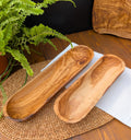 Two olive wood bread baskets showing natural grain, set on a woven placemat beside a potted green fern.