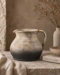 A rustic terracotta jug vase with a textured, distressed finish, featuring a gradient from light beige at the top to a dark charcoal grey at the base. It is styled on a stone ledge next to a linen cloth, a book, and a dried flower vase.