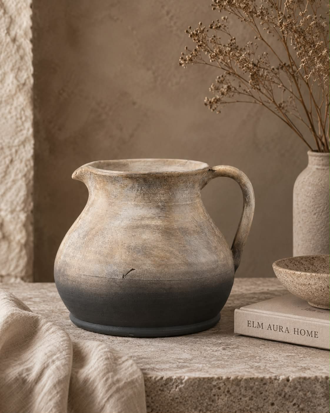 A rustic terracotta jug vase with a textured, distressed finish, featuring a gradient from light beige at the top to a dark charcoal grey at the base. It is styled on a stone ledge next to a linen cloth, a book, and a dried flower vase.