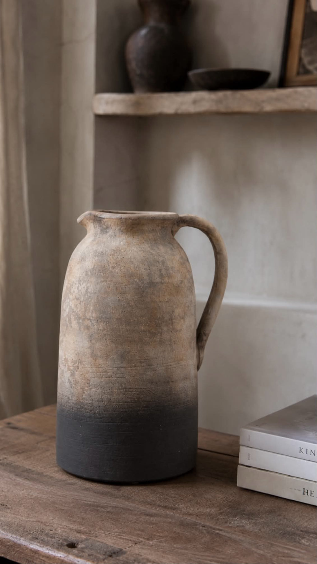 A rustic terracotta jug vase with a natural earthy finish, featuring a gradient from light tan at the top to a dark charcoal gray at the base, sitting on a wooden table next to a stack of books.