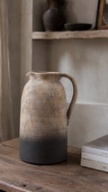 A rustic terracotta jug vase with a natural earthy finish, featuring a gradient from light tan at the top to a dark charcoal gray at the base, sitting on a wooden table next to a stack of books.