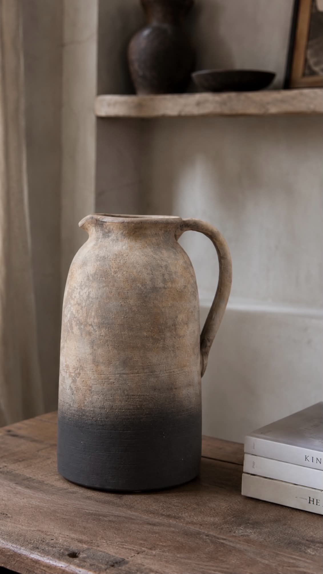 A rustic terracotta jug vase with a natural earthy finish, featuring a gradient from light tan at the top to a dark charcoal gray at the base, sitting on a wooden table next to a stack of books.