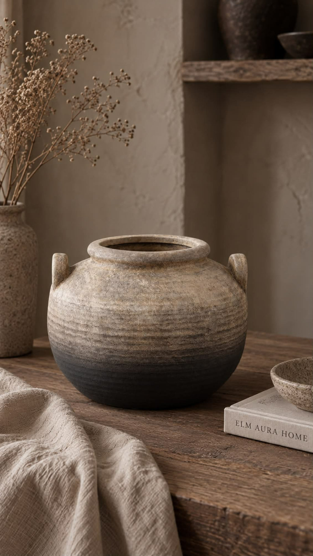 A rustic terracotta vase with a two-tone gradient from beige to dark charcoal, styled on a wooden table with linen fabric and dried flowers in a warm, earthy room setting.
