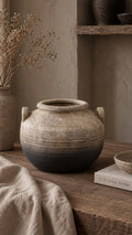 A rustic terracotta vase with a two-tone gradient from beige to dark charcoal, styled on a wooden table with linen fabric and dried flowers in a warm, earthy room setting.