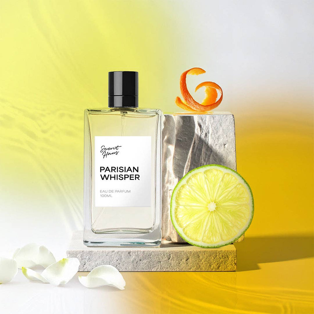 A perfume bottle labeled Parisian Whisper Eau de Parfum 100ml, styled with a lime slice, an orange peel curl, and white rose petals on a reflective yellow and white surface.
