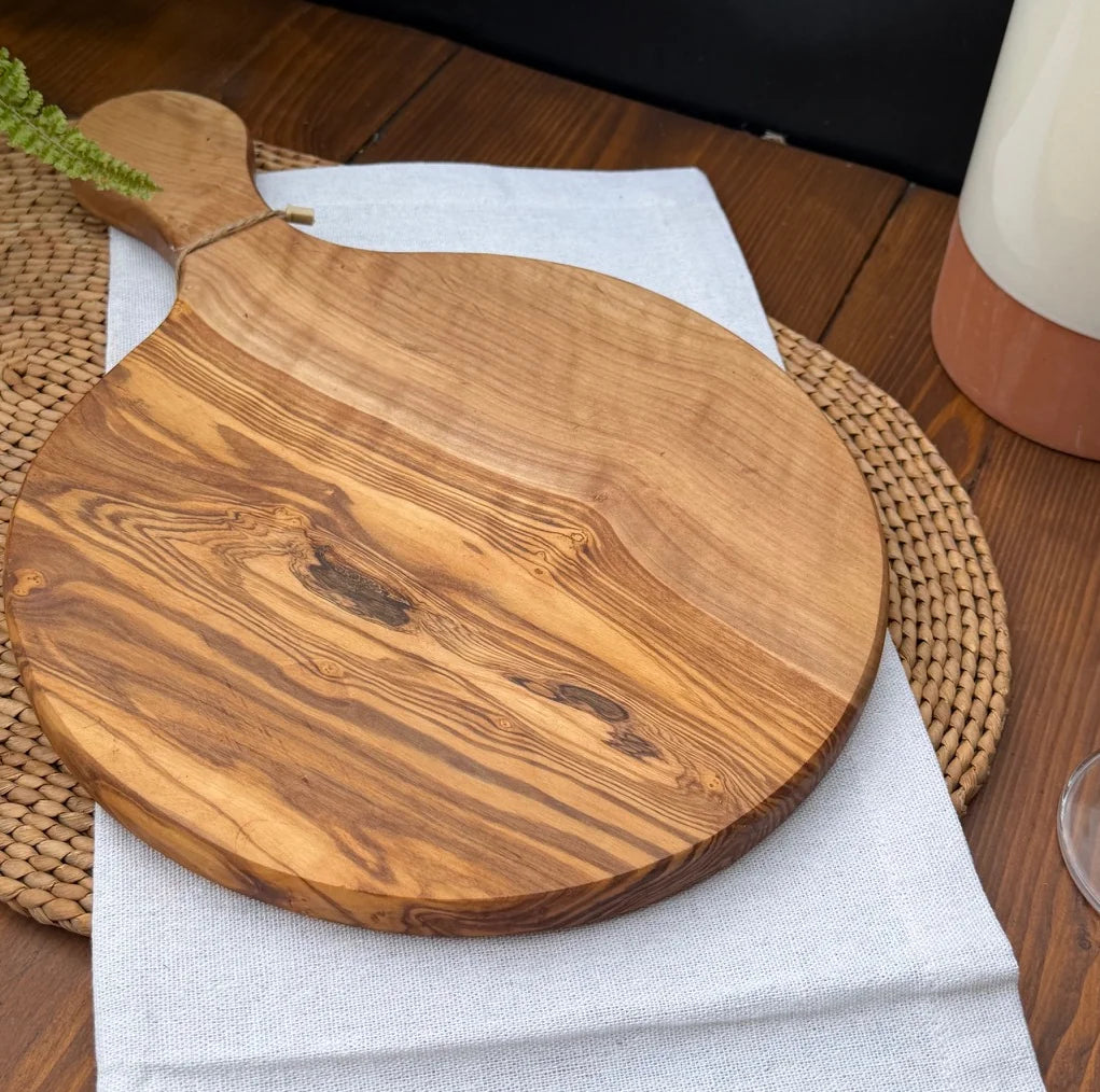 Round olive wood serving board with handle on a white linen napkin over a woven placemat.
