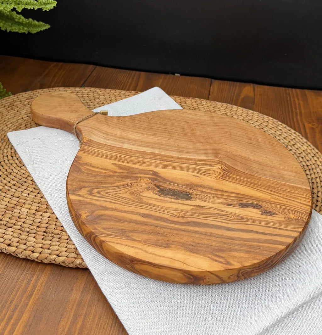 Round olive wood serving board with handle resting on a white cloth over a woven placemat.