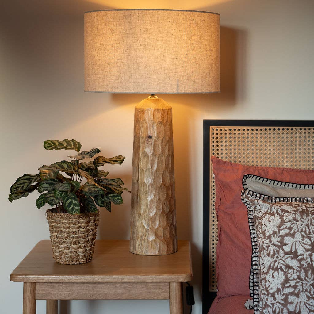 Hand-carved wooden table lamp with a linen shade placed on a side table next to a bed with soft pillows.