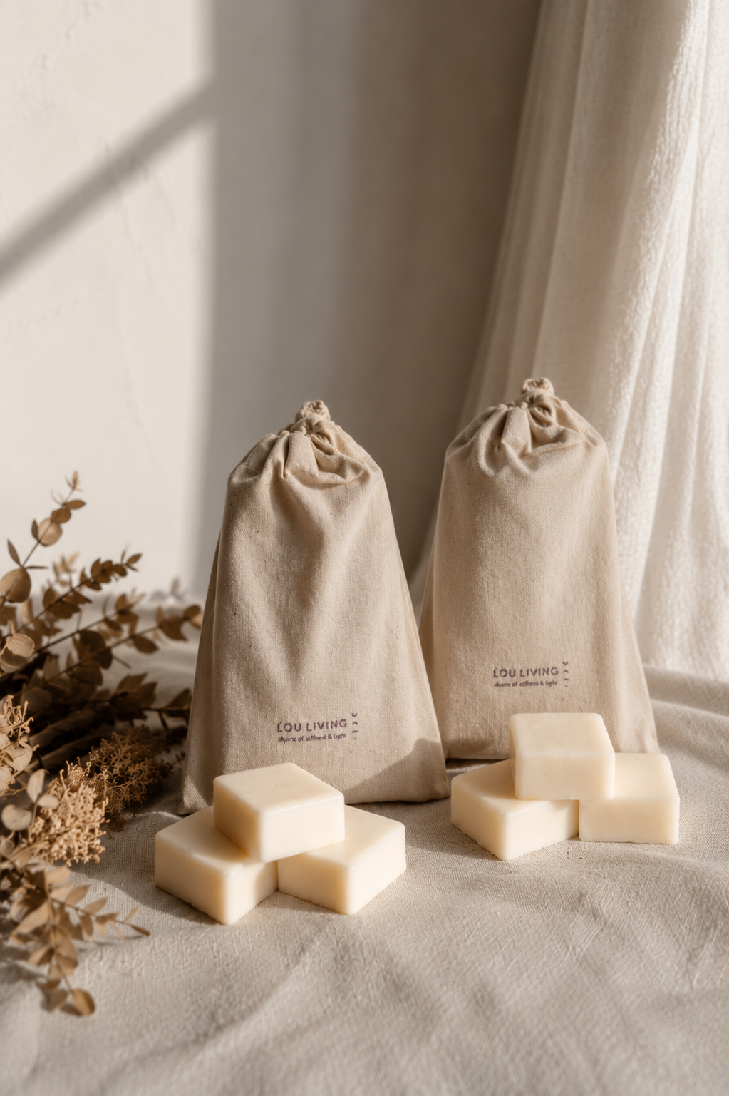 Two beige drawstring pouches arranged on a draped fabric surface with several pale cream Still Light natural essential oil wax melts around them and dried floral decor at the side.