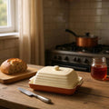 Terracotta butter dish with a butter knife and fresh bread on a solid wooden kitchen table.
