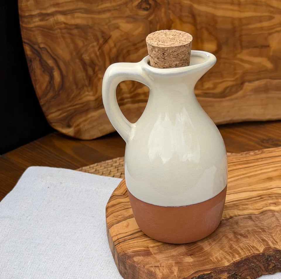 Terracotta oil drizzler with glossy off-white glaze and cork stopper on a wooden serving board.