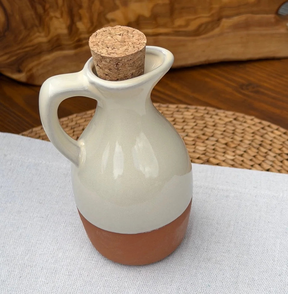 Terracotta oil drizzler with handle, glossy off-white glaze, and cork stopper on a light fabric surface.