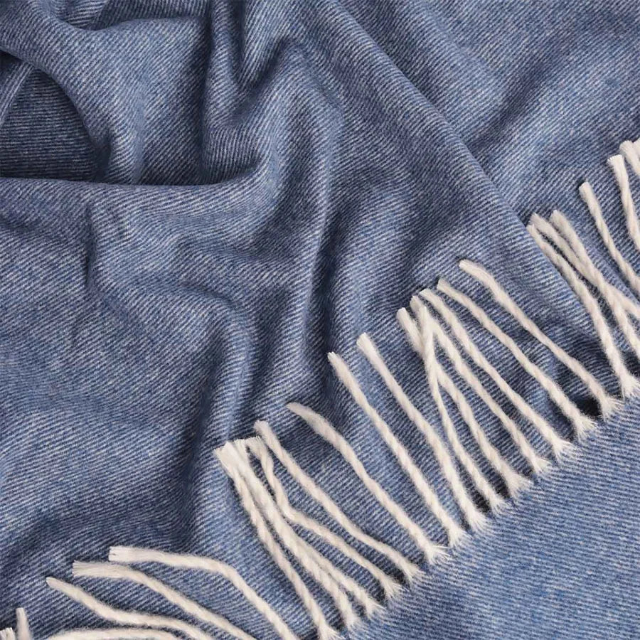 A close-up of a blue polo cashmere and wool throw blanket with off-white fringe along one edge, showing the fabric texture and folds.