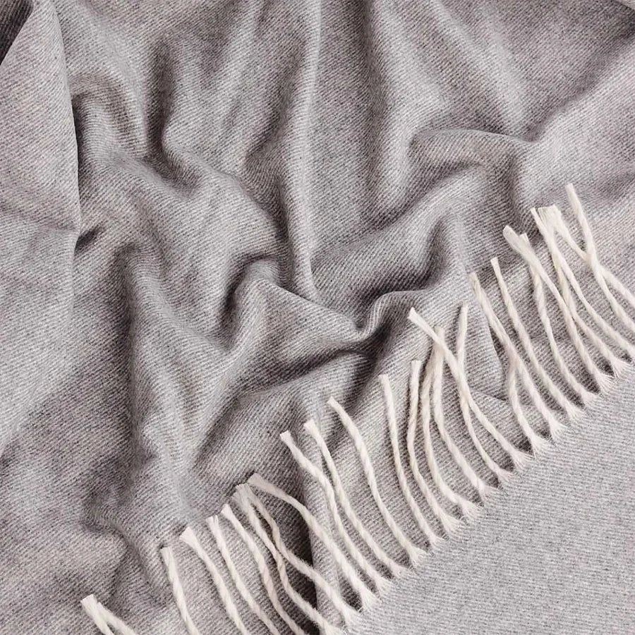 A close-up of a soft grey cashmere and wool throw blanket with off-white fringe along one edge, draped in gentle folds.