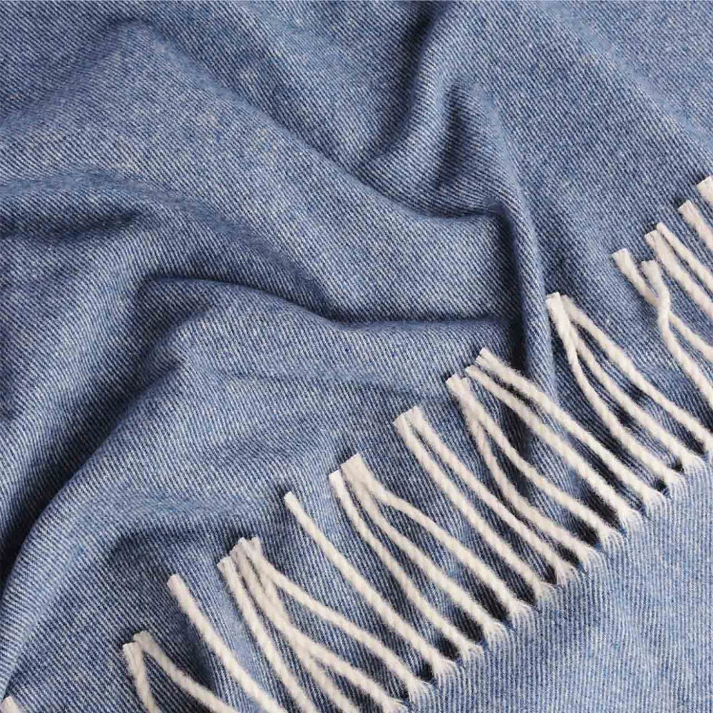 A close-up of a blue Steppe merino throw blanket with cream fringe along one edge, showing the fabric texture and folds.