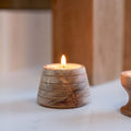 A small lit tea light in a textured, ribbed wooden holder with a warm flame, set on a light surface with a softly blurred wooden background.