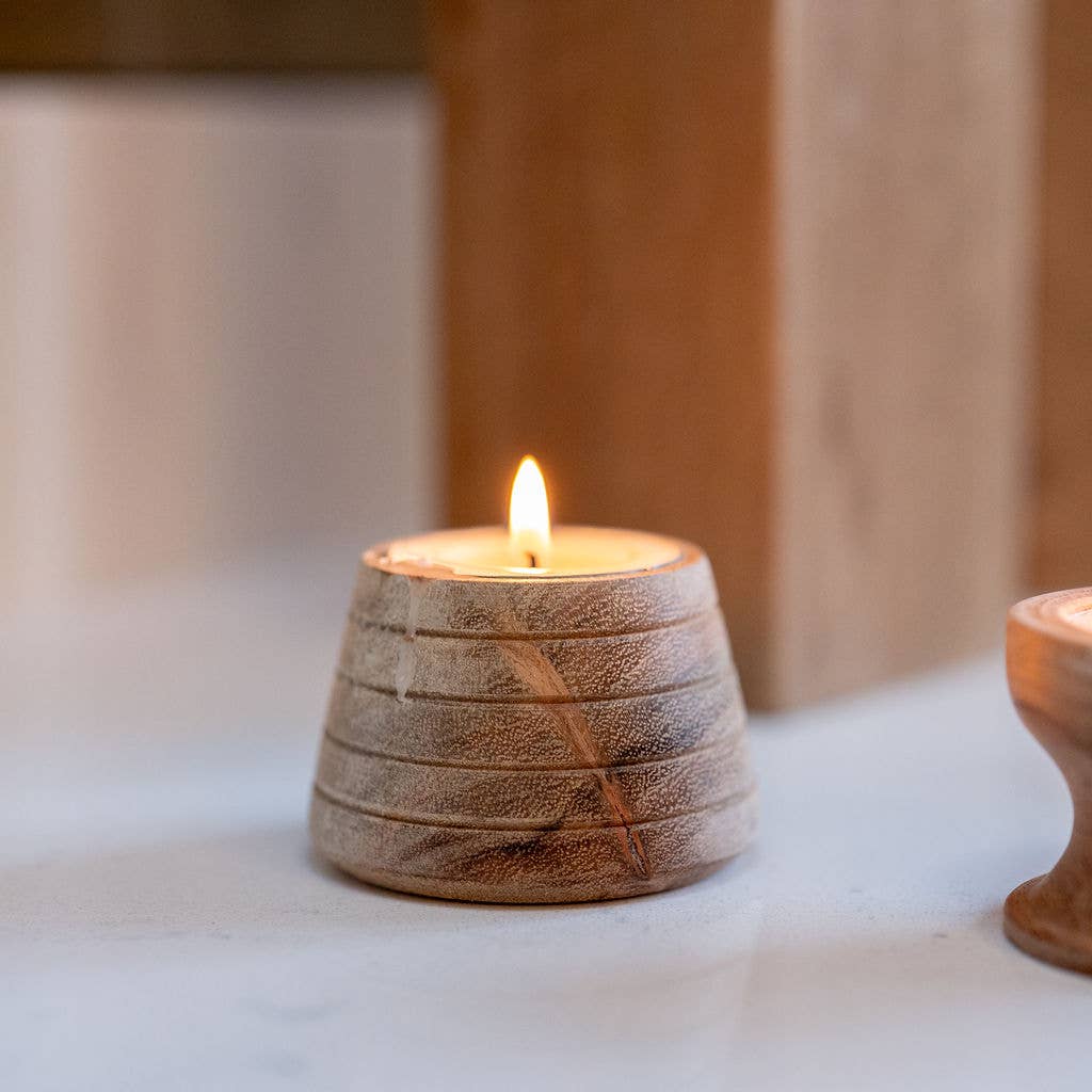 A small lit tea light in a textured, ribbed wooden holder with a warm flame, set on a light surface with a softly blurred wooden background.