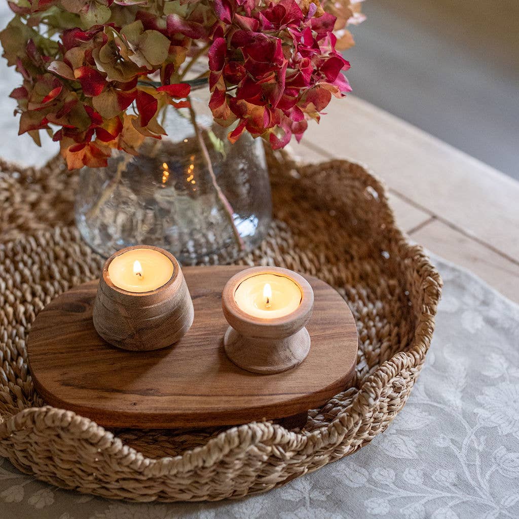 Two lit tea light holders on a small oval wooden tray, placed inside a woven round tray with dried flowers in a glass vase in the background.