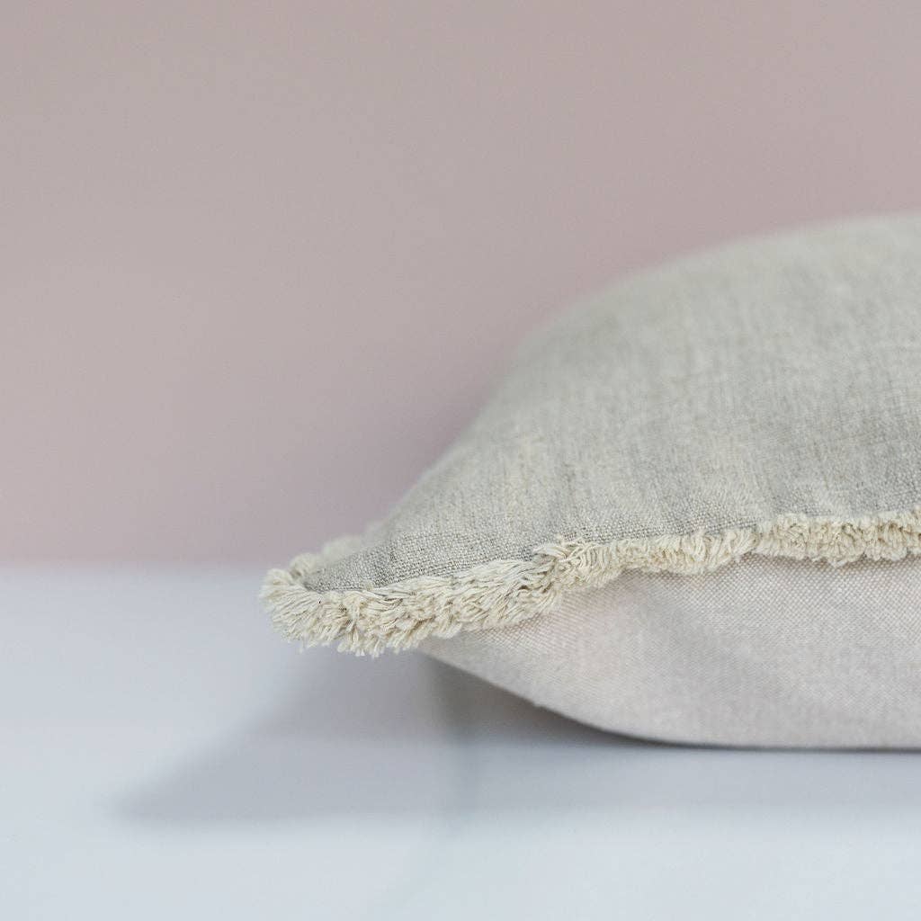 Close-up of a textured beige linen cushion cover with decorative fringing on a muted pink background.