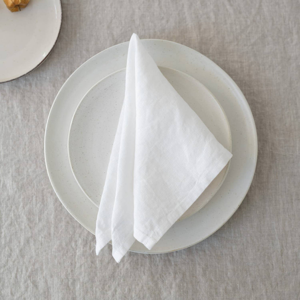 A folded white linen napkin placed on a white ceramic plate resting on a textured linen tablecloth.