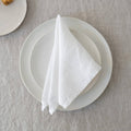 A folded white linen napkin placed on a white ceramic plate resting on a textured linen tablecloth.