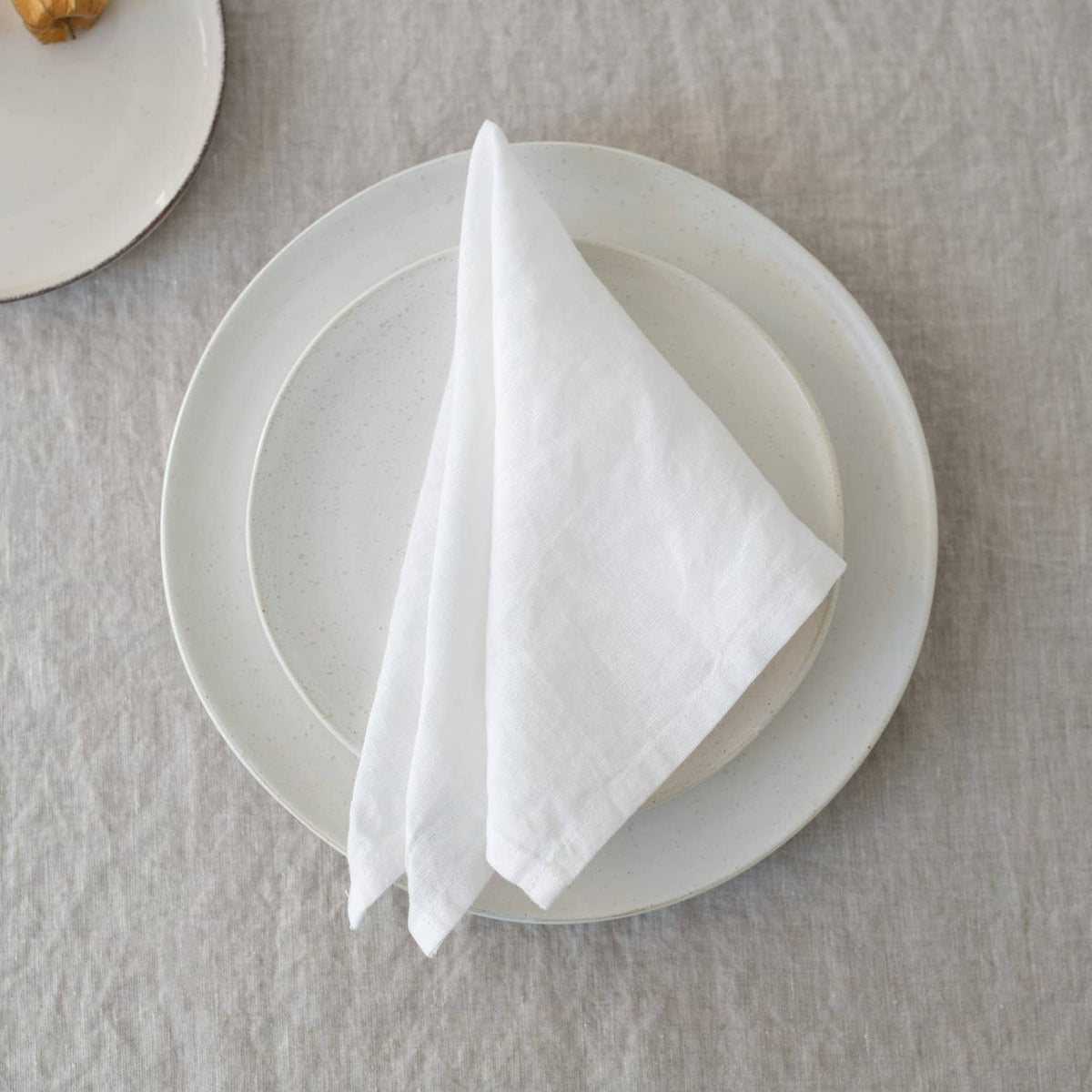 A folded white linen napkin placed on a white ceramic plate resting on a textured linen tablecloth.