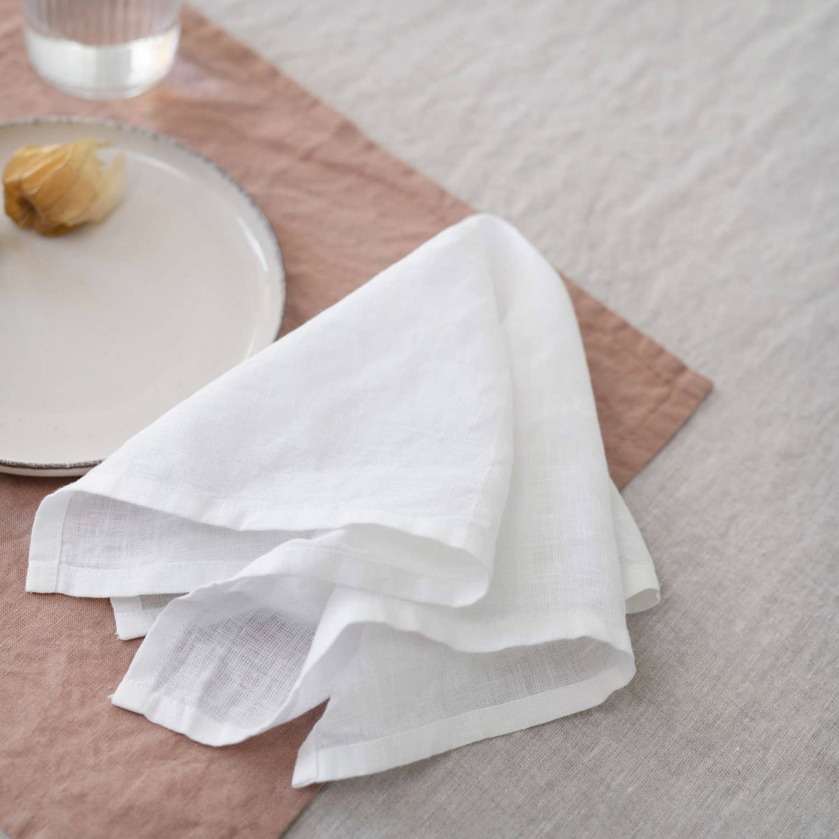 White linen napkin with a subtle woven texture styled on a dusty pink placemat over a light beige linen tablecloth, with a ceramic plate and glassware in the background.