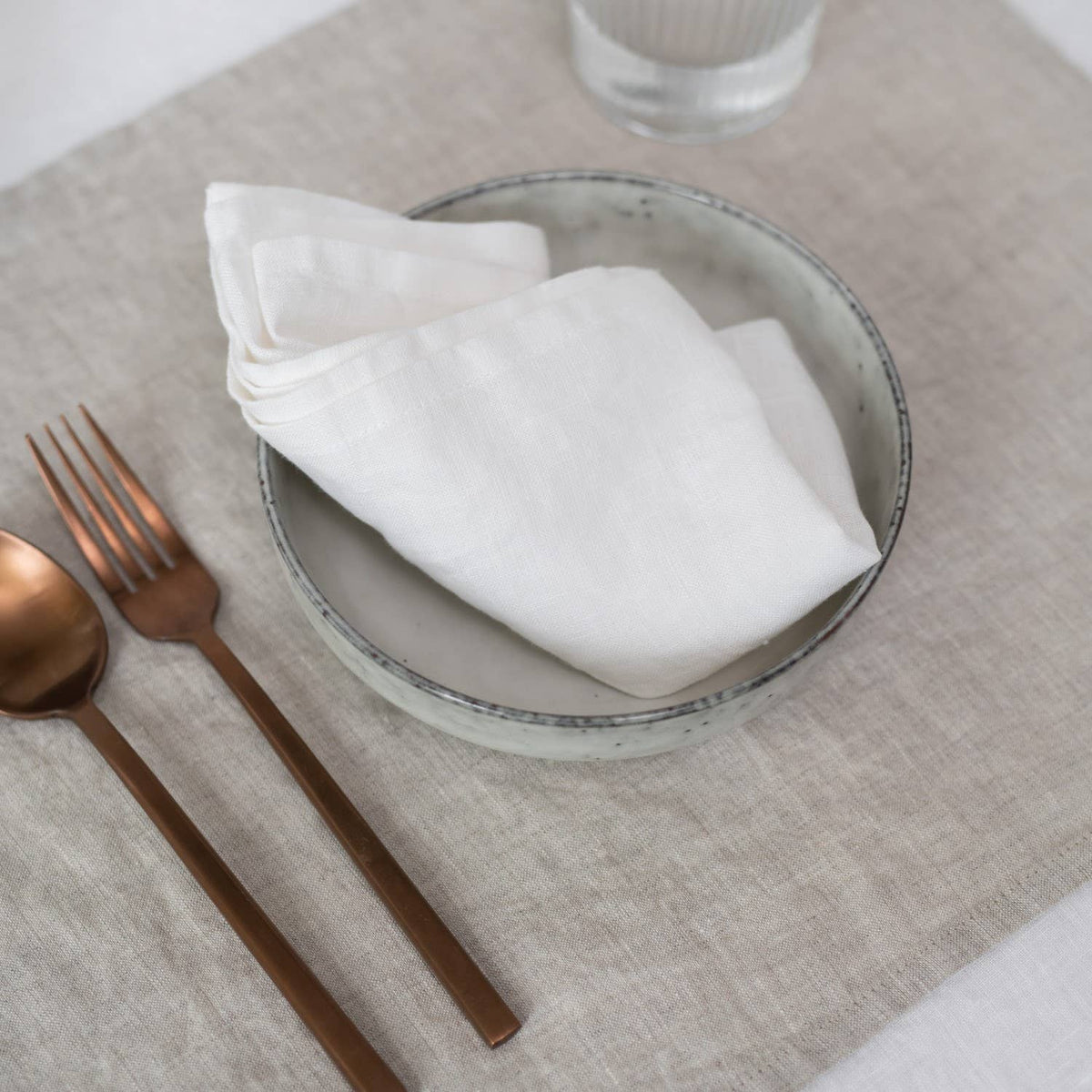 A white linen napkin placed inside a ceramic bowl on a dusty pink placemat, set with a fork and spoon alongside.
