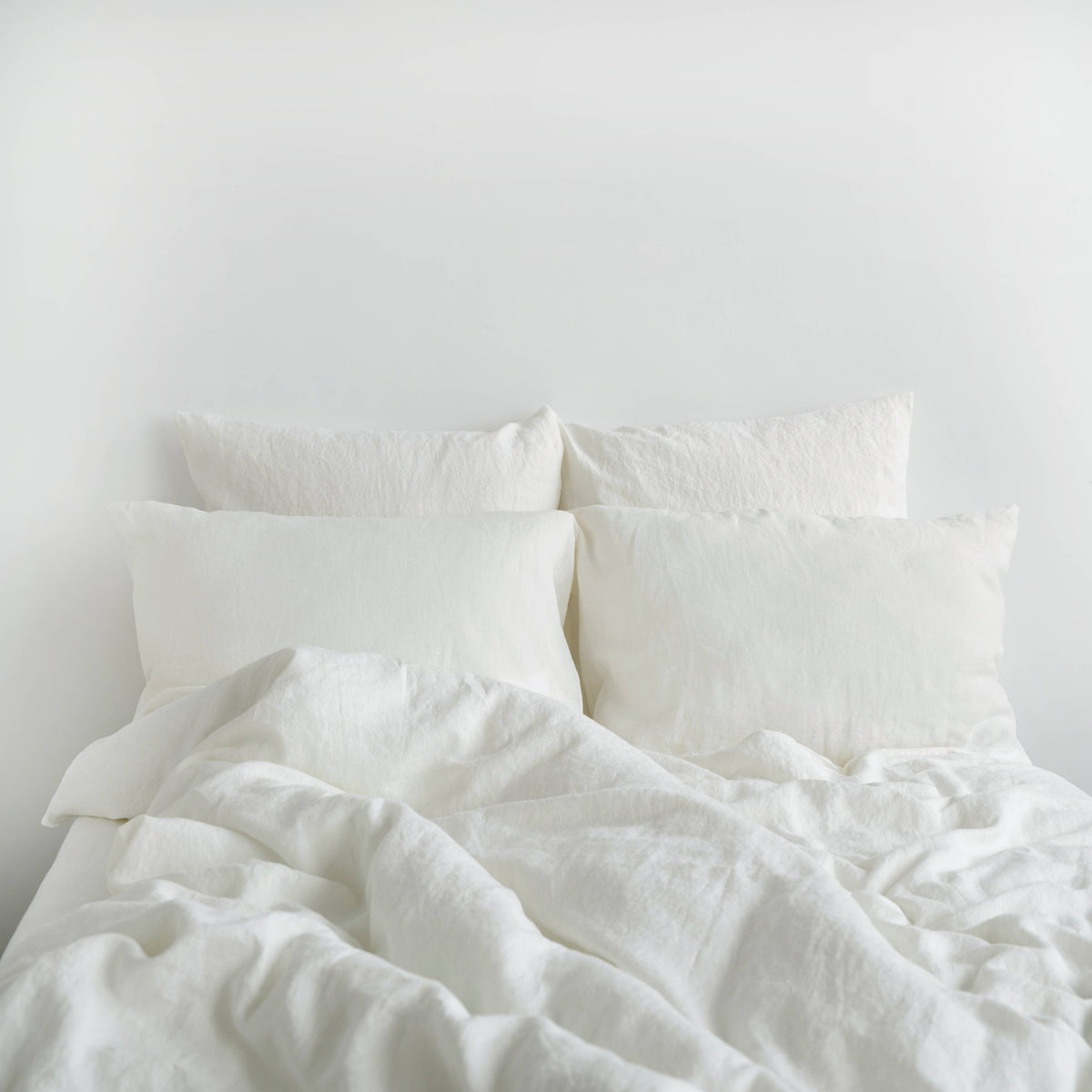 White linen bedding set featuring a relaxed, rumpled duvet and multiple pillows arranged on a bed against a plain white wall.
